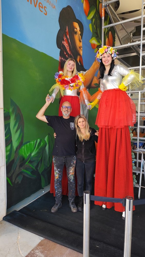 O artista Fly Pontes e a sua companheira a tirar uma foto com duas jovens em andas, em frente ao mural finalizado "Mostra da Laranja 2025". A presença das andas ajuda a perceber a escala e grandeza (5m×6m) da obra em Silves.