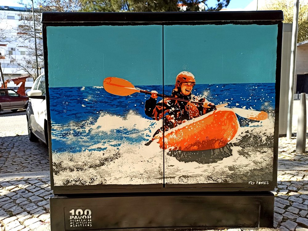 Pintura "A Alegria e a Adrenalina da Canoagem" terminado num armário de electricidade em Albufeira. Uma figura feminina desce uma onda num caiaque, pintada em tons vibrantes de laranja contra o azul do oceano. Técnica Stencil e Spray.