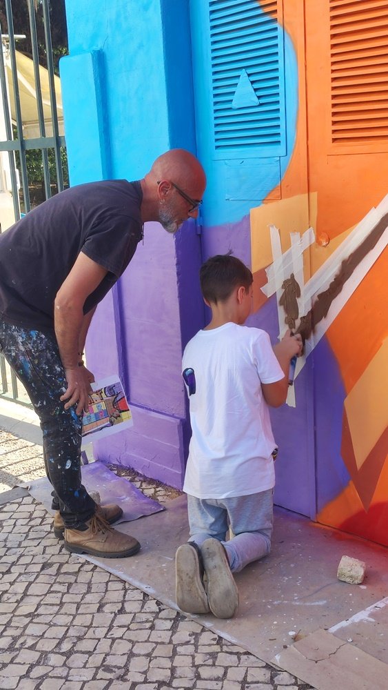 Um dos alunos de artes a pintar uma das linhas do mural Pop Art. O aluno usa fita cola (masking) para garantir que as linhas ficam direitas e precisas. O artista Fly Pontes supervisiona o trabalho no Posto de Transformação (PT).