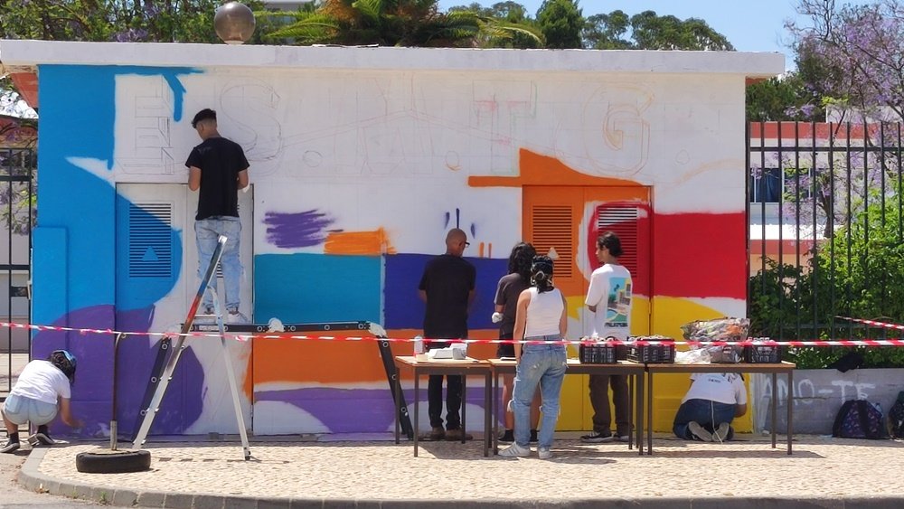 Alunos da Escola Secundária Manuel Teixeira Gomes a pintar o fundo do mural Pop Art no Posto de Transformação (PT). Grande parte do fundo já está pintado e alunos aprendem a técnica de uso da lata de spray. Colaboração na arte urbana.