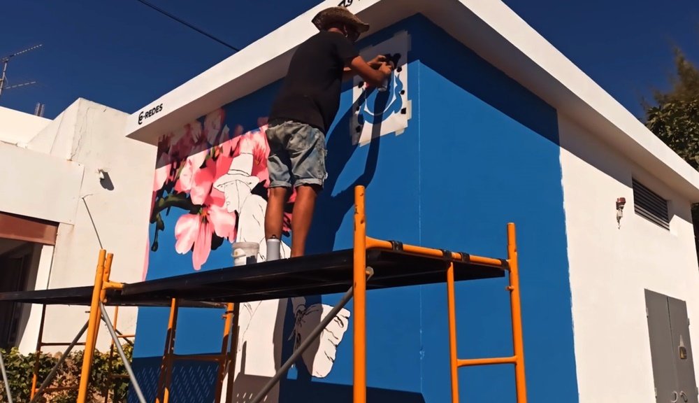 O artista Fly Pontes a pintar o stencil do logotipo da Junta de Freguesia de Almancil. O mural "Homenagem à Marquinhas" está completo, e o logotipo marca o apoio institucional ao projecto de arte urbana.