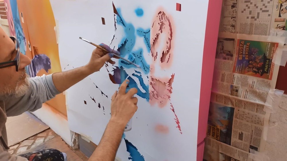 O artista Fly Pontes a aplicar uma das camadas do stencil de um dos gatos do mural "Gatinhos Pop". O outro gato já é visível no armário de electricidade. O artista usa um pauzinho para pressionar a folha, garantindo que a tinta não passe por baixo do stencil e não estrague a nitidez da obra.