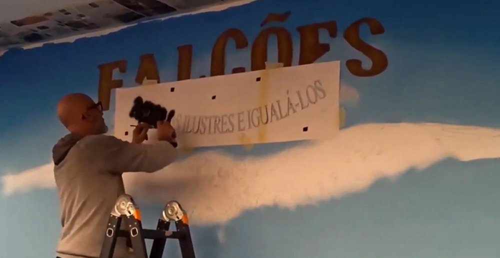 O artista Fly Pontes em cima do escadote, a aplicar o stencil do lema "Imitar os Ilustres e Igualá-los". O artista usa spray à base de água na pintura do mural "Os Falcões" da BA11 em Beja.