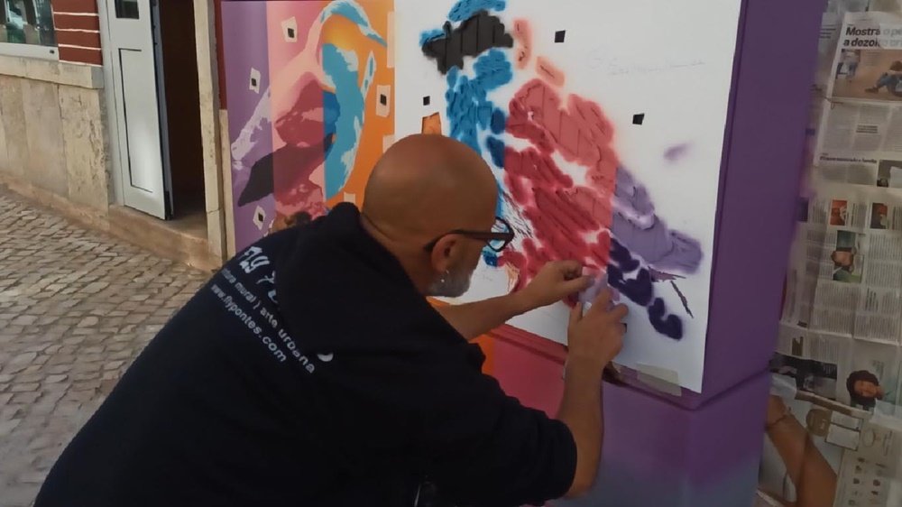 O artista Fly Pontes a aplicar outra camada de stencil. A vista mais afastada permite ver a progressão do mural e as cores (azul, rosa, violeta) já aplicadas no outro pardal. Foco na técnica de stencil multicamadas e na composição geral.