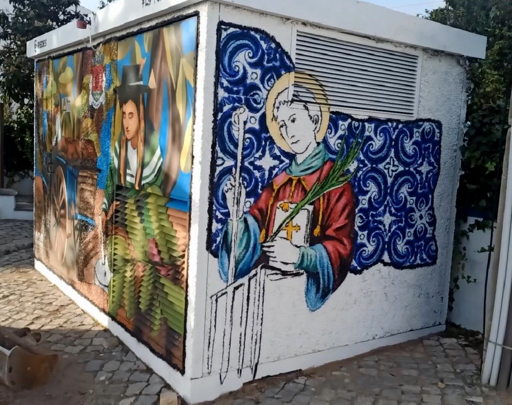 Mural lateral do PT em Almancil (Fé e História). A pintura homenageia o patrono S. Lourenço, incorporando o design dos azulejos da igreja local, criando uma ponte visual entre fé e arte no espaço público.