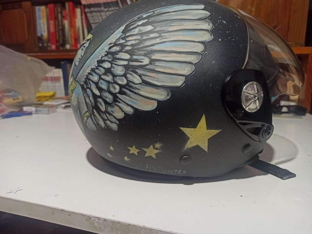 Close-up da lateral do capacete personalizado, mostrando os detalhes das estrelas amarelas em progressão e o fundo esfumado com salpicos de tinta. Ilustra a mistura de Spray e Marcadores usada na técnica.