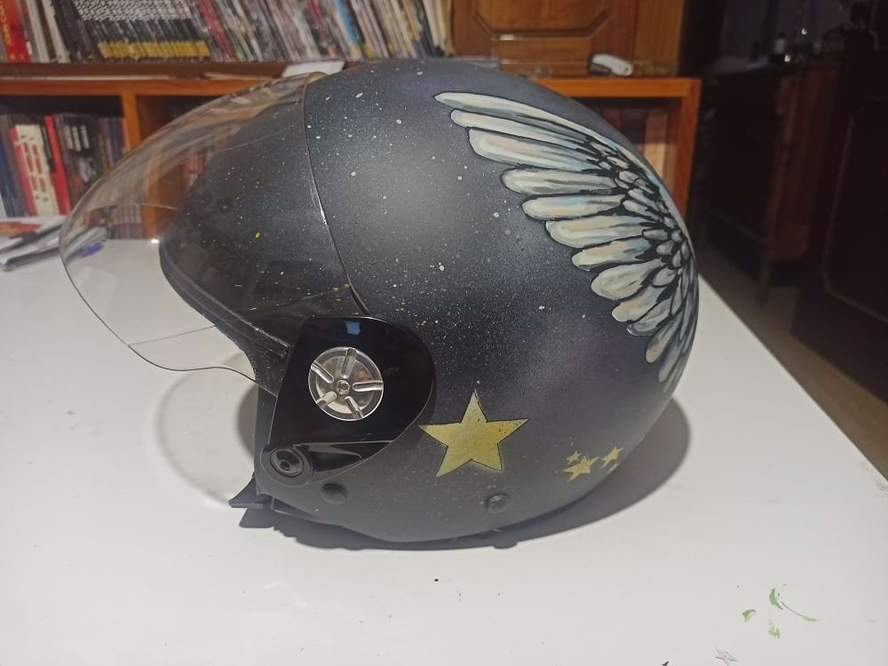 Close-up da lateral do capacete personalizado, mostrando os detalhes das estrelas amarelas em progressão e o fundo esfumado com salpicos de tinta. Ilustra a mistura de Spray e Marcadores usada na técnica.