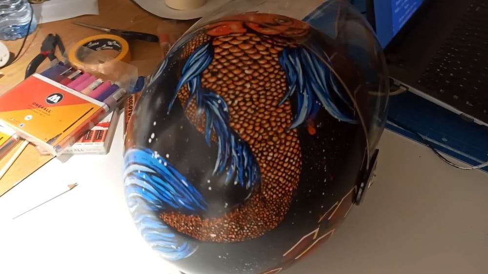 Capacete de mota personalizado com o design de uma Carpa Koi vibrante. O fundo foi criado com spray acrílico e a figura central detalhada com marcadores Molotow One4all, garantindo cores ricas e durabilidade.