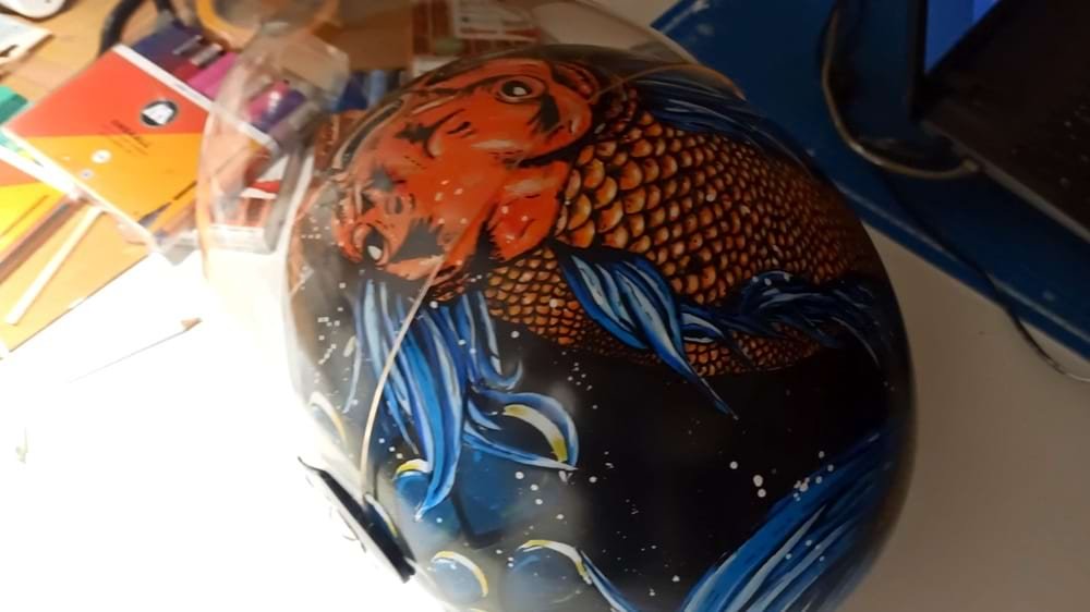 Capacete de mota personalizado com o design de uma Carpa Koi vibrante. O fundo foi criado com spray acrílico e a figura central detalhada com marcadores Molotow One4all, garantindo cores ricas e durabilidade.