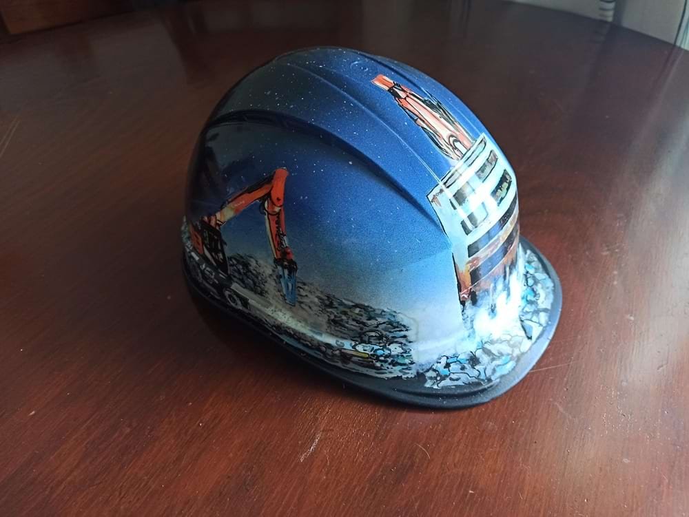 Detalhe da lateral do capacete de obra, mostrando a maquinaria pesada específica desenhada para o cliente. A imagem demonstra a precisão dos marcadores na representação técnica.