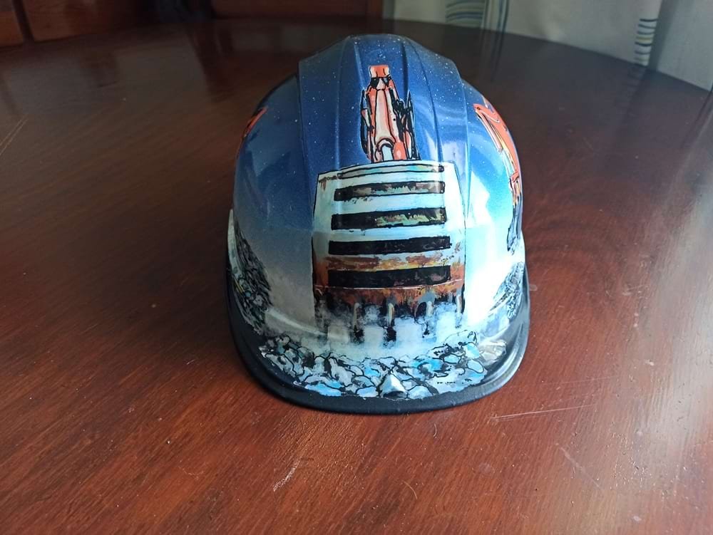 Capacete de obra personalizado com o tema "Obra e Maquinaria". A frente é dominada pela pá de uma retroescavadora. A pintura foi executada com marcadores One4all e spray, e protegida com verniz acrílico para durabilidade no trabalho.
