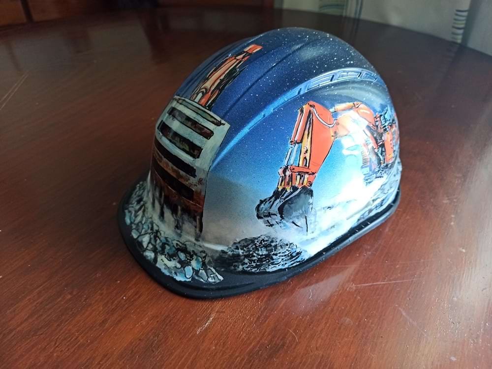 Detalhe da lateral do capacete de obra, mostrando a maquinaria pesada específica desenhada para o cliente. A imagem demonstra a precisão dos marcadores na representação técnica.