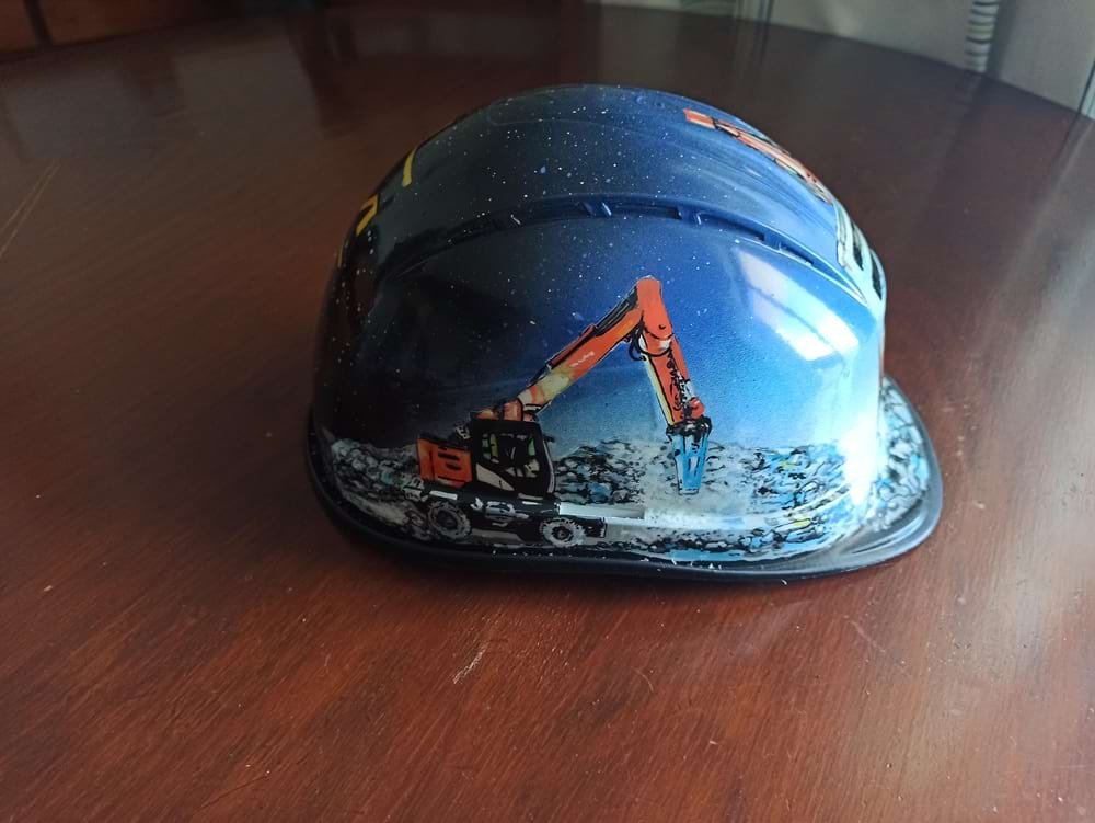 Detalhe da lateral do capacete de obra, mostrando a maquinaria pesada específica desenhada para o cliente. A imagem demonstra a precisão dos marcadores na representação técnica.