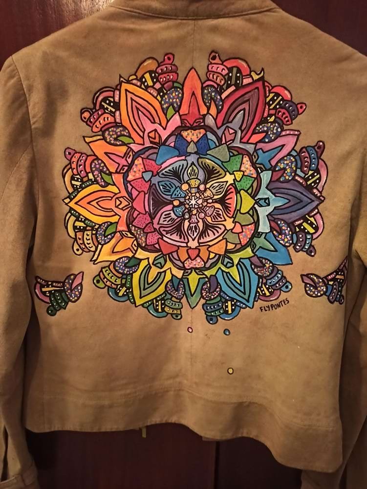Mandala pintada à mão no casaco de camurça, mostrando a profusão de cores e os degradés ricos em tons de azul, laranja, rosa e verde. É visível o rigor do detalhe da pintura.