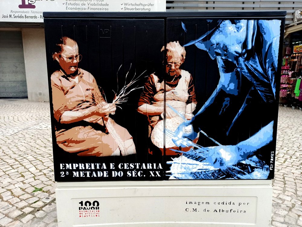 Pintura em Albufeira que homenageia a cestaria e a empreita. Um homem em azul tece um cesto (primeiro plano), e duas mulheres trabalham na empreita ao fundo, recriando o trabalho manual e comunitário da região.