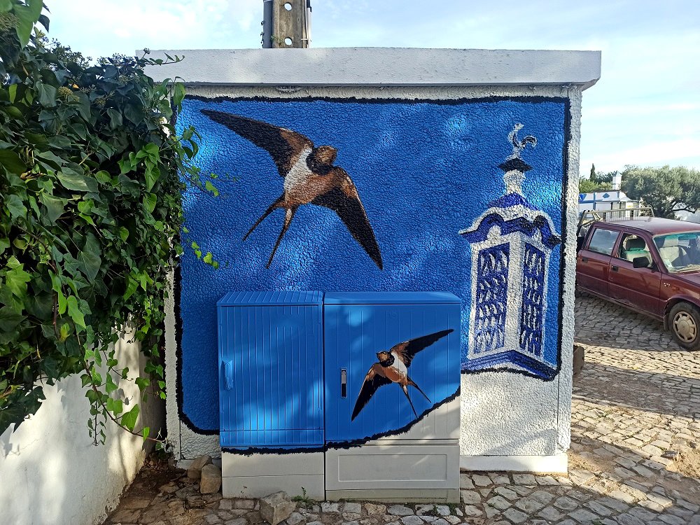Mural lateral do PT em Almancil (Tradição e Natureza). A obra apresenta uma chaminé centenária e andorinhas a voar, pintadas em Stencil e Spray sobre um fundo todo azul para criar forte contraste.