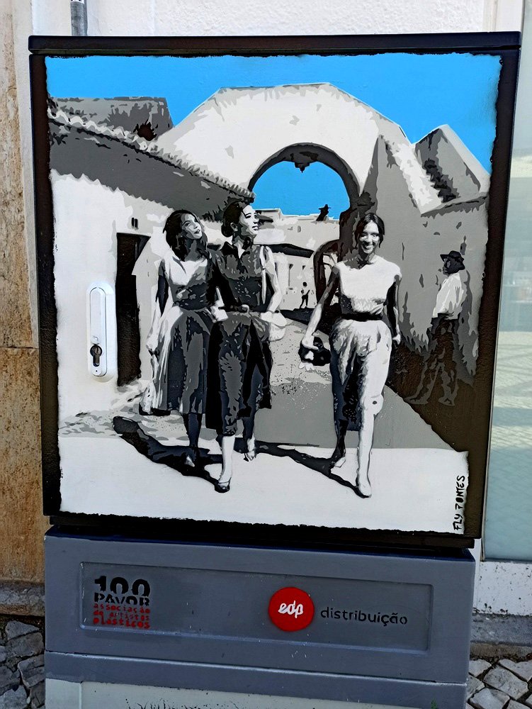 Pintura "Choque Cultural do Turismo" em Albufeira. A cena, incluindo três mulheres e observadores, está em tons de cinza (passado histórico), contrastando com o céu azul vibrante (o futuro). A obra recria o nascimento do turismo na Rua do Arco.