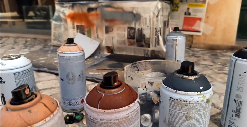 Close-up das latas de spray de diferentes cores, com os bicos (caps) já colocados, e um recipiente separado para organizar os bicos. Destaque para as ferramentas essenciais da técnica stencil e spray.