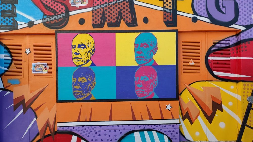 Grande plano do retrato de Manuel Teixeira Gomes pintado no Posto de Transformação (PT). A pintura segue a técnica de Andy Warhol (múltiplas imagens e cores vivas). Uma folha de referência está colada no PT, e a homenagem Pop Art está quase terminada.