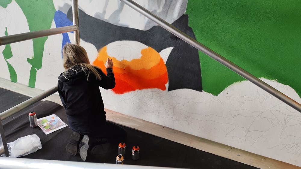 Colaboração na fase final do mural da Mostra da Laranja em Silves. O artista recebe a ajuda da sua companheira, a pintar as laranjas na parte de baixo da obra. Destaque para o trabalho de equipa e os toques finais nos frutos.