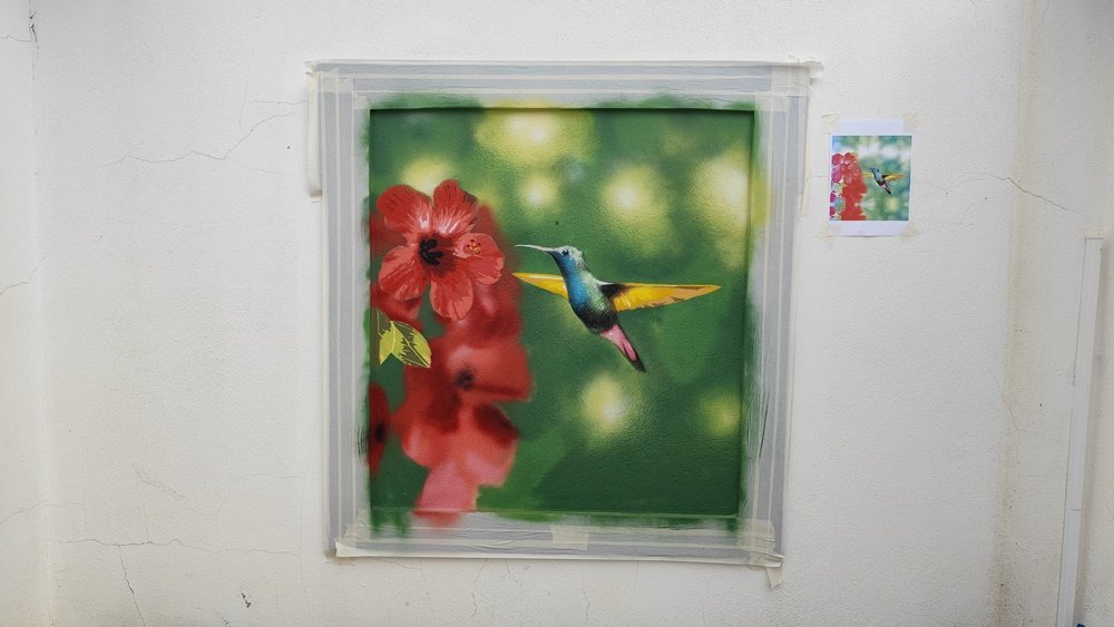 A pintura 'Colibri na Janela Falsa' já sem a máscara, mostrando o contraste entre o fundo desfocado e o colibri realista. Uma folha de referência é visível na parede ao lado da pintura. Arte em stencil e spray.