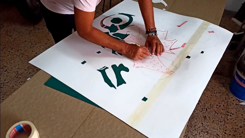 O artista Fly Pontes a cortar manualmente um stencil da motorizada 50cc para o mural "O Espírito da Estrada". Vêem-se as partes já cortadas e as linhas vermelhas a indicar as áreas a preservar. Precisão na técnica de stencil.