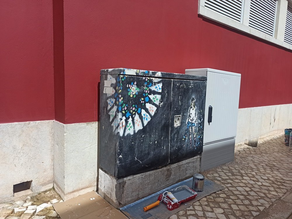 Armário de electricidade em S. B. Messines com a pintura anterior (reprodução da Sé de Évora e uma turista) em estado de degradação. Foto do antes, mostrando a necessidade de revitalização do mobiliário urbano.