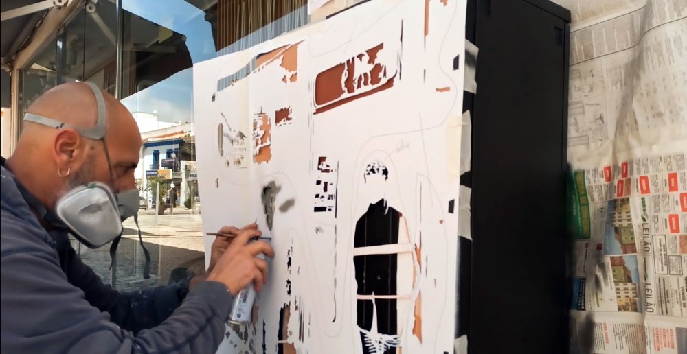 O artista Fly Pontes a pintar uma das camadas de stencil com uma lata de spray, enquanto segura o pauzinho para fixar o molde. Este método garante a precisão e nitidez dos detalhes da pintura da Primeira Farmácia.