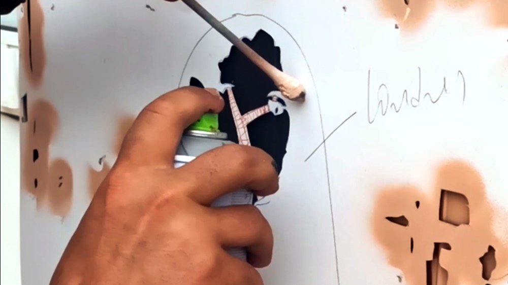 Detalhe da aplicação de uma camada do stencil. A inscrição "Londres" na folha de stencil funciona como um guia para a cor correcta a ser aplicada com o spray. Um pauzinho segura a folha para maior precisão.