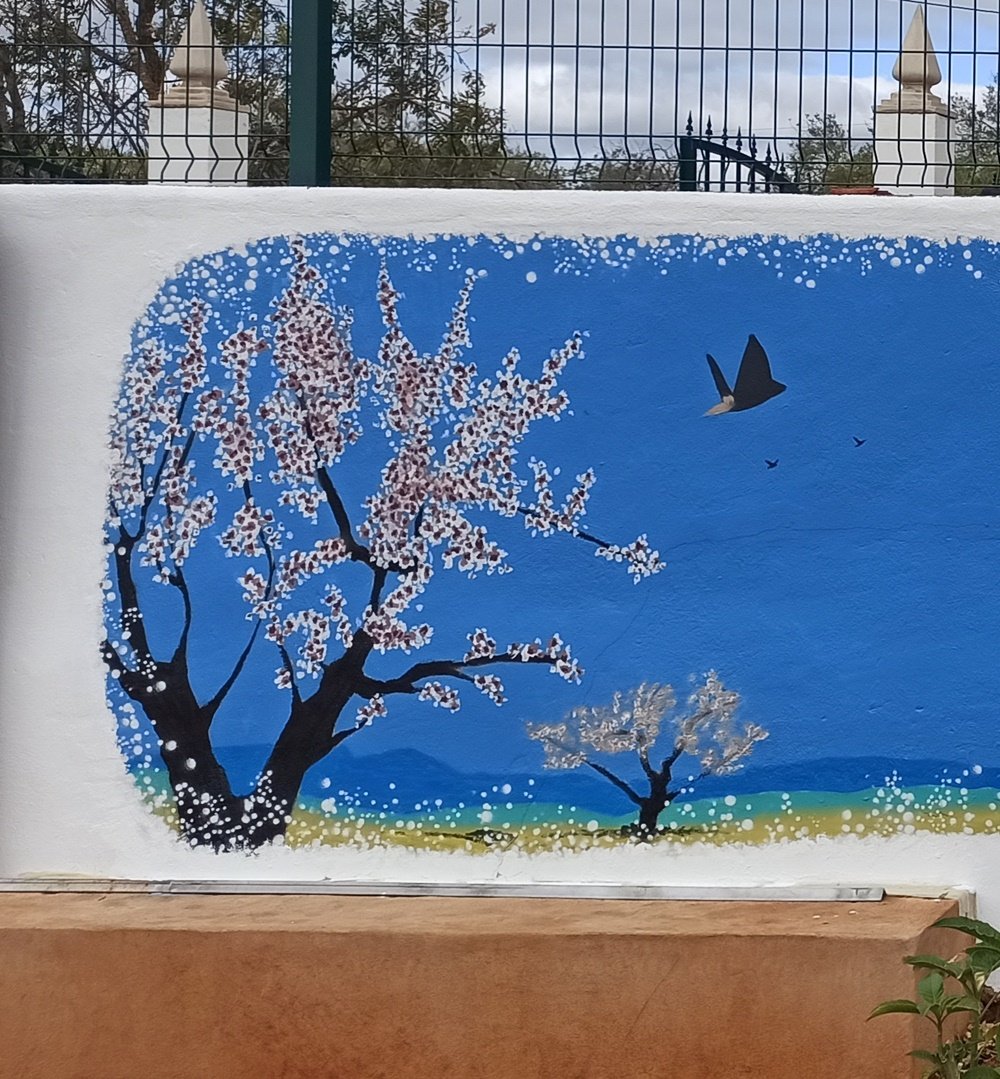 Detalhe de uma amendoeira em flor (estilo japonês) em primeiro plano, com um pardal em silhueta a levantar vôo. A composição inclui outra amendoeira ao longe, criando profundidade sob o céu azul.