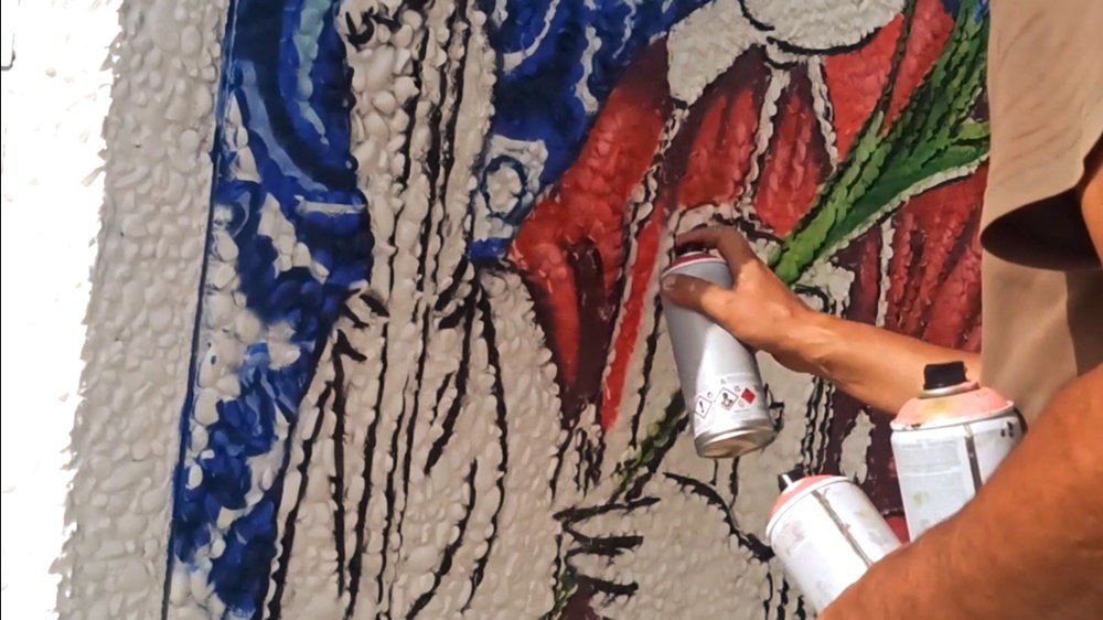 Processo de pintura do mural de S. Lourenço (Lateral do PT em Almancil). O artista Fly Pontes utiliza uma lata de spray para aplicar os tons vermelhos da vestimenta da figura, tendo mais latas de outras cores no braço esquerdo.