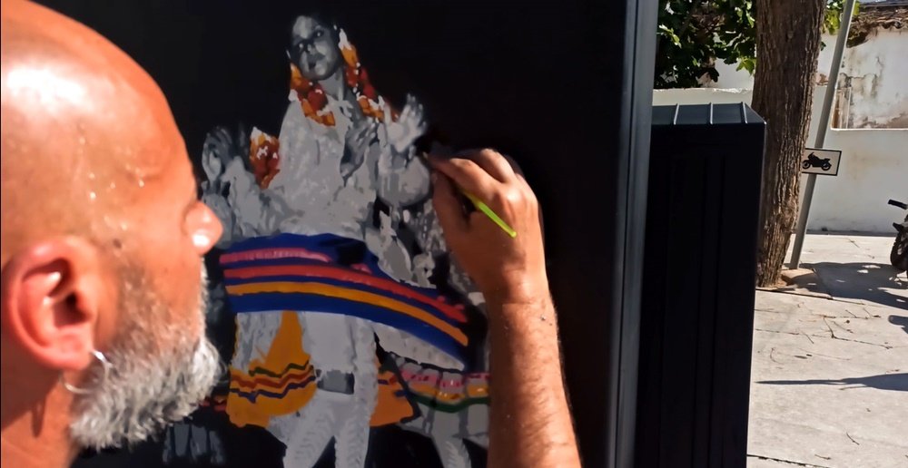 Vista aproximada do artista Fly Pontes a segurar um pincel e a dar os retoques finais na pintura. A figura feminina do rancho folclórico já está bem definida, mostrando a atenção aos detalhes para o acabamento.