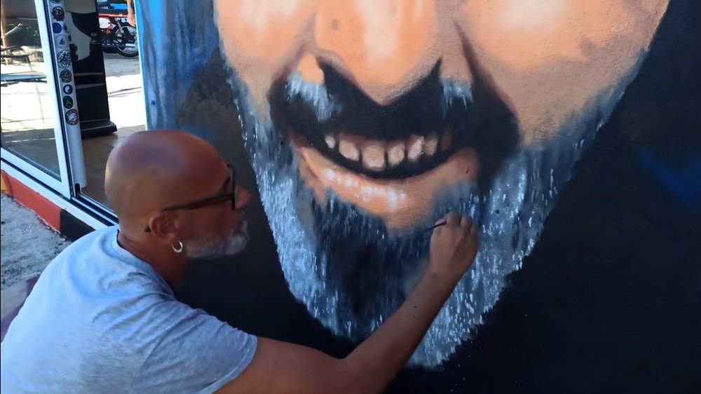 Grande plano do retrato de Nelson Francês. O artista Fly Pontes usa um pincel fino para pintar os pormenores dos pelos da barba, adicionando realismo e textura à figura principal do mural VEOH.
