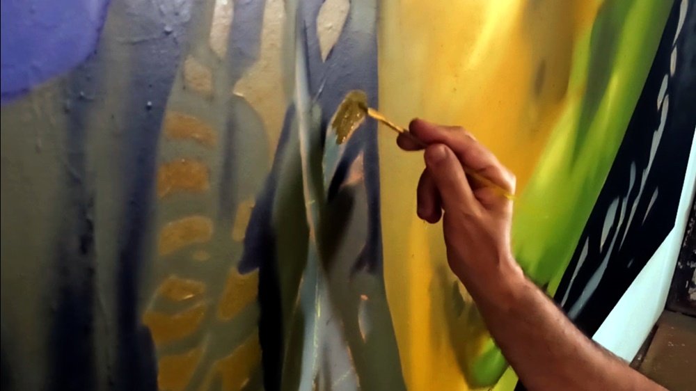 Close-up do artista Fly Pontes a pintar os detalhes da roupa do retrato, utilizando um pincel fino. A mudança de ferramenta demonstra a necessidade de precisão e acabamento minucioso nas áreas que complementam a figura.