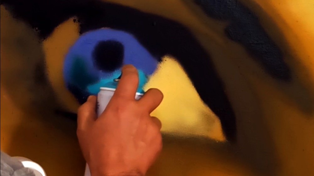 Close-up do artista Fly Pontes a pintar os olhos em tons azuis com spray no retrato do mural "Estudo de Cor e Escala". A aplicação de azul nos olhos cria um ponto focal e um contraste intencional dentro da figura em tons de verde.
