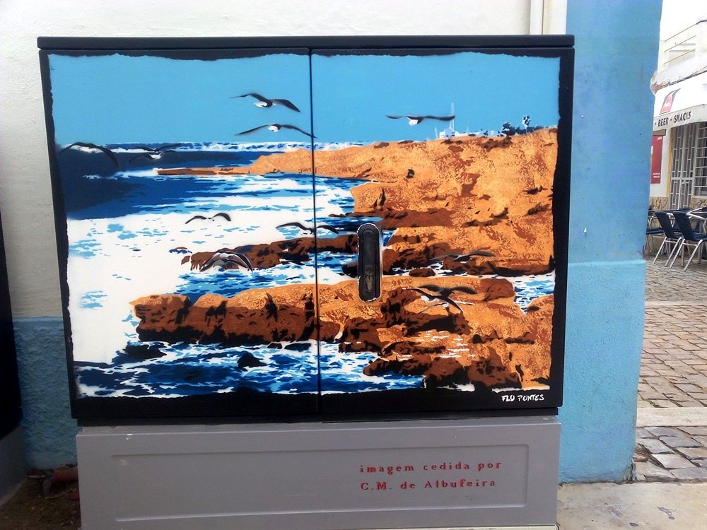 Pintura "As Falésias da Costa de Albufeira" terminado num armário de electricidade. As falésias estão pintadas em tons quentes de castanho e o mar em azul vibrante, com ondas em rebentação. A cena inclui gaivotas no ar. Técnica Stencil e Spray.