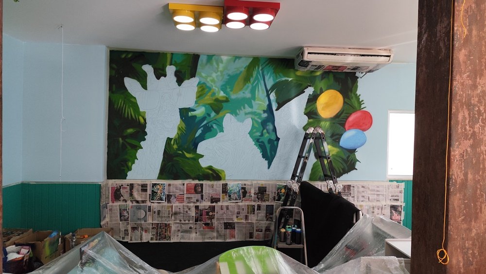 A selva de fundo do mural está pronta, mostrando profundidade com verdes escuros, médios e claros próximos e azuis esverdeados distantes. Os três balões que o macaco segura também estão pintados. Arte urbana em progresso no "Mural da Selva" em Albufeira.
