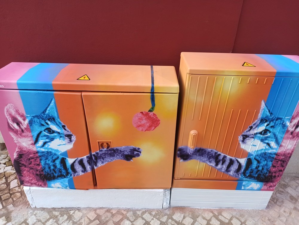 Pintura "Gatinhos Pop" finalizado, com vista de cima. É visível o fio do novelo que continua até à parte de trás do armário e os sinais de aviso (Perigo de Morte) que são integrados na obra. Arte urbana em mobiliário eléctrico em Messines.