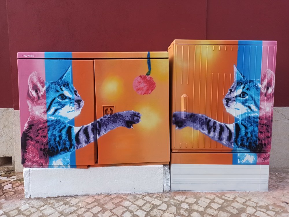Mural em miniatura "Gatinhos e o Novelo" terminado, transformando um armário de electricidade em S. Bartolomeu de Messines. A obra usa a técnica Stencil e Spray com cores vibrantes (fundo laranja, gatos em azuis/violetas/rosas).