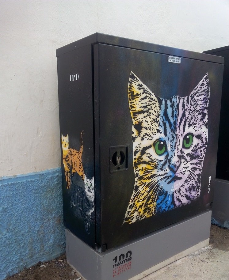 Vista lateral/frontal do armário de electricidade com a pintura. Na frente, o Gato Pop. Na lateral, dois gatos (um amarelo e outro cinzento) com os rabos levantados, olhando para cima, em posição de esperar comida. Todos os gatos criados com a técnica de stencil.