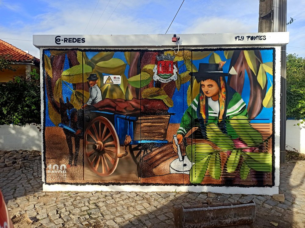 Mural da frente do PT em Almancil (Projecto Tradição, Fé e Património Agrícola). A pintura homenageia a alfarroba e a tradição agrícola, com uma algarvia a moer o fruto, uma carroça antiga e folhas de alfarroba em macro.