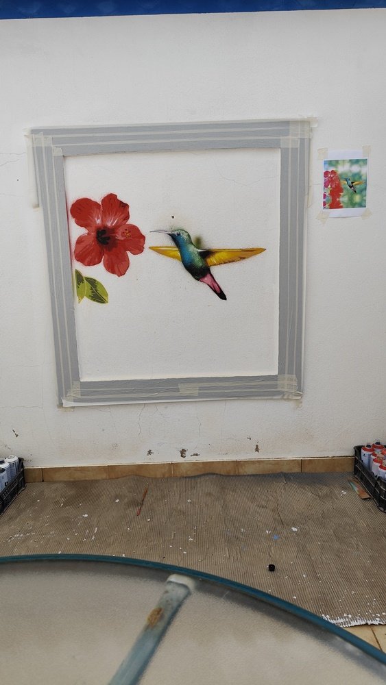 Início da pintura do 'Colibri na Janela Falsa'. O colibri e a flor vermelha mais próxima já visíveis, pintados com técnica de stencil e spray. Arte urbana em progresso.