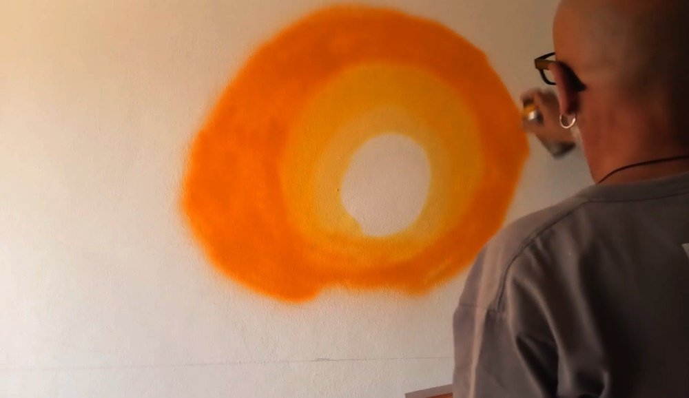 O artista Fly Pontes a começar a pintar o pôr do sol do mural "O Pôr do Sol do Pescador", aplicando os primeiros tons de laranja. Esta fase define a atmosfera quente do céu algarvio, que servirá de fundo para a silhueta.