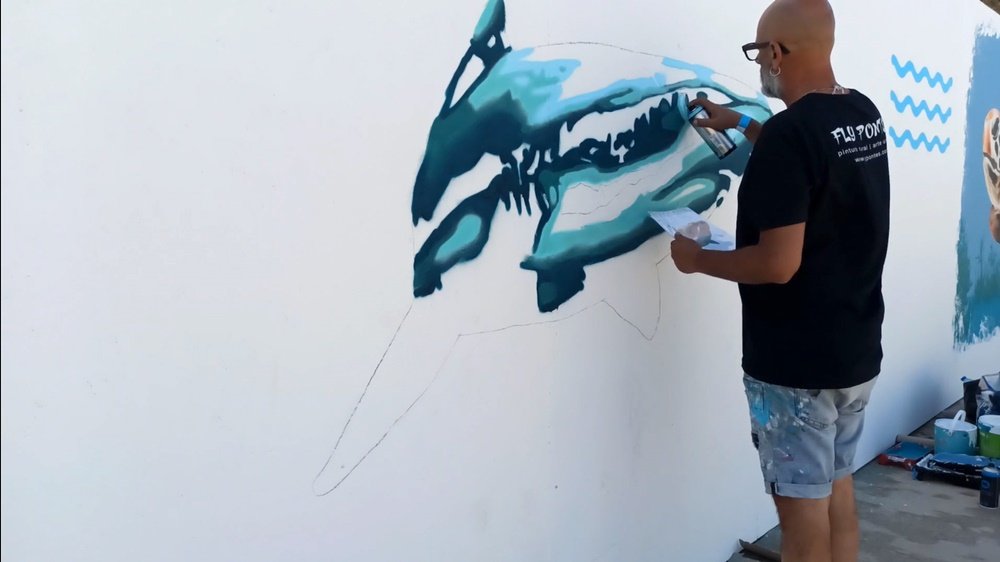 Início da pintura do Tubarão-Branco no Festival Mar Me Quer, em Portimão. O mural está a ser pintado em tons azuis, e o artista tem na mão uma folha de referência. Registo da criação de arte urbana de conservação marinha.