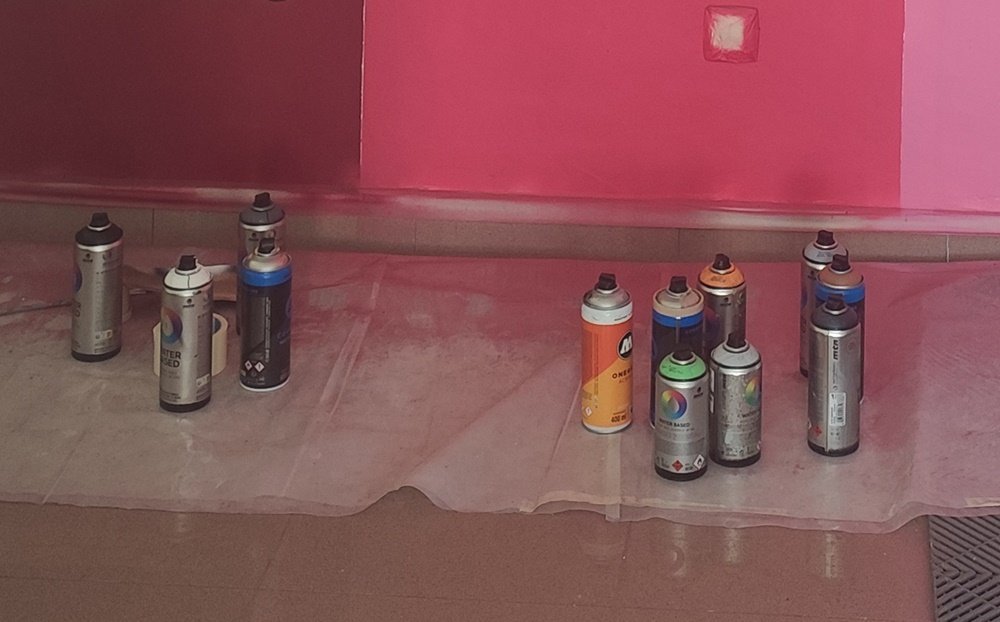 Vista em plano médio de latas de spray à base de água (water based) no chão do local da pintura, durante o mural da Algarve Paint Store. Foco nas ferramentas e na escolha de material sustentável.