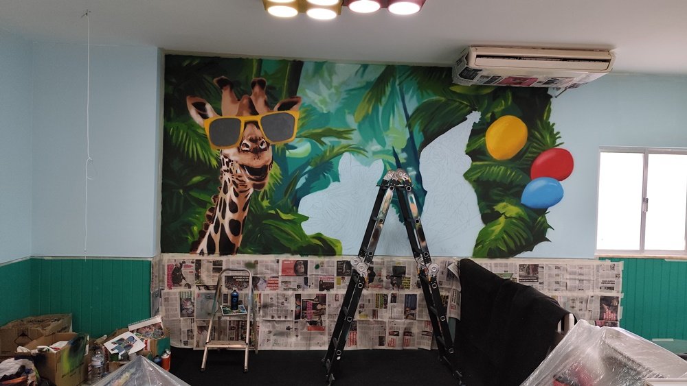 Girafa do Mural da Selva (4x2,8m) pintada. A girafa usa óculos com lentes que seriam posteriormente corrigidas para dar realismo e profundidade. Foco no detalhe técnico do spray no "Eat, Learn and Play" em Albufeira.