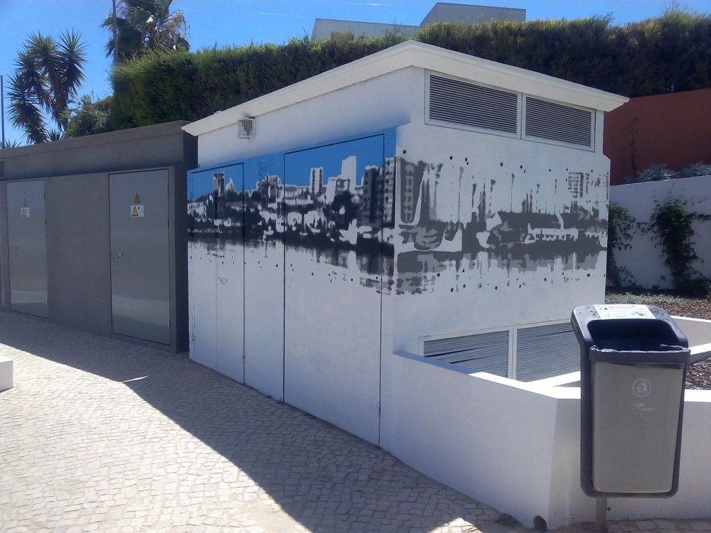 Posto de Transformação com duas fases de pintura. Um lado mostra o skyline com o céu azul concluído, enquanto o outro lado tem apenas o skyline em cinza (sem céu). São visíveis as marcações e fitas usadas para o stencil multicamadas no mural.