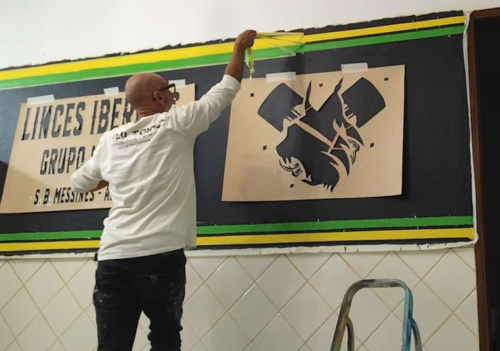 Artista a remover fita-cola das linhas rectas amarelo e verde no mural do bar. Stencils do lince e letras visíveis.