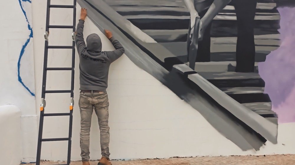 O artista Fly Pontes ao longe, de costas, a esticar um fio de marcação para desenhar as linhas do caminho de ferro direitas. A pintura do trabalhador do caminho de ferro é visível. A imagem realça a grande escala do mural na Estação Almancil - Neche (Esteval).