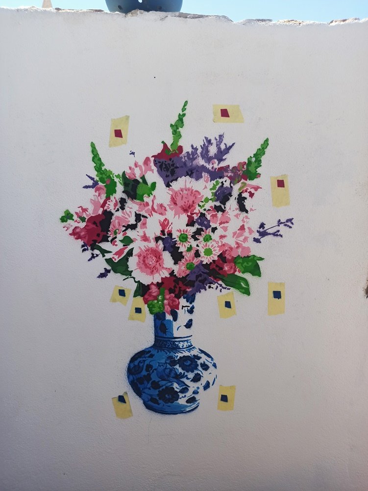 A pintura "Natureza Morta em Stencil" a meio da pintura. Vêem-se várias cores aplicadas nas flores (rosas, vermelhos, verdes) e as marcações na parede para o posicionamento preciso dos stencils multicamadas.